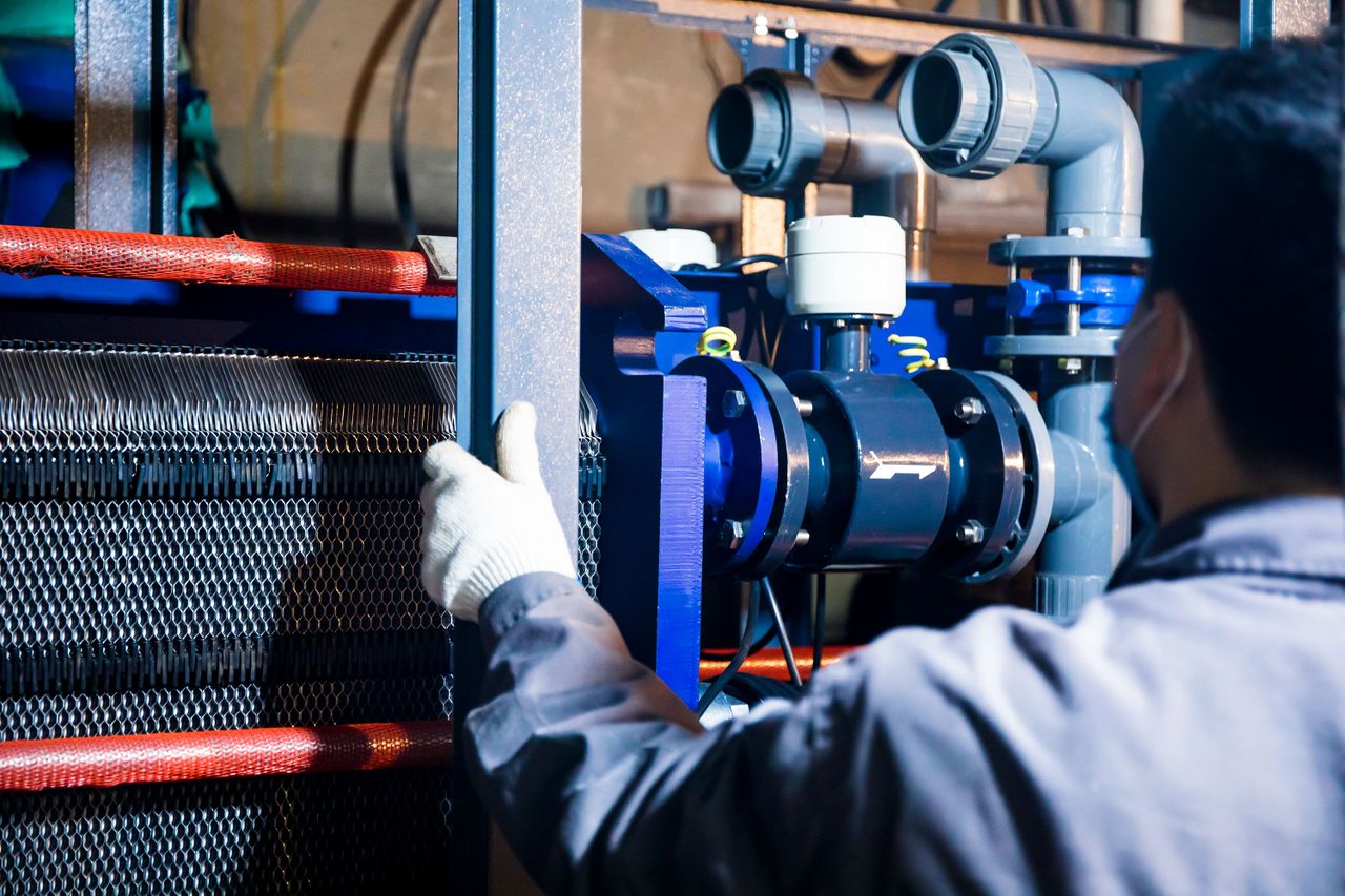 our-story A worker in blue sleeves inspects machinery pipes and equipment at an industrial plant.